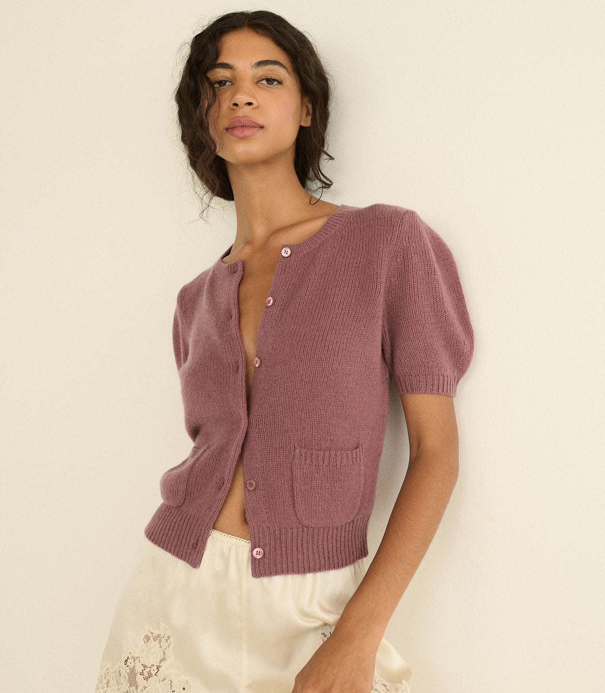 A woman wearing a short-sleeve knit cardigan in a dusty mauve-pink color, paired with white-colored shorts, posing against a plain off-white background.
