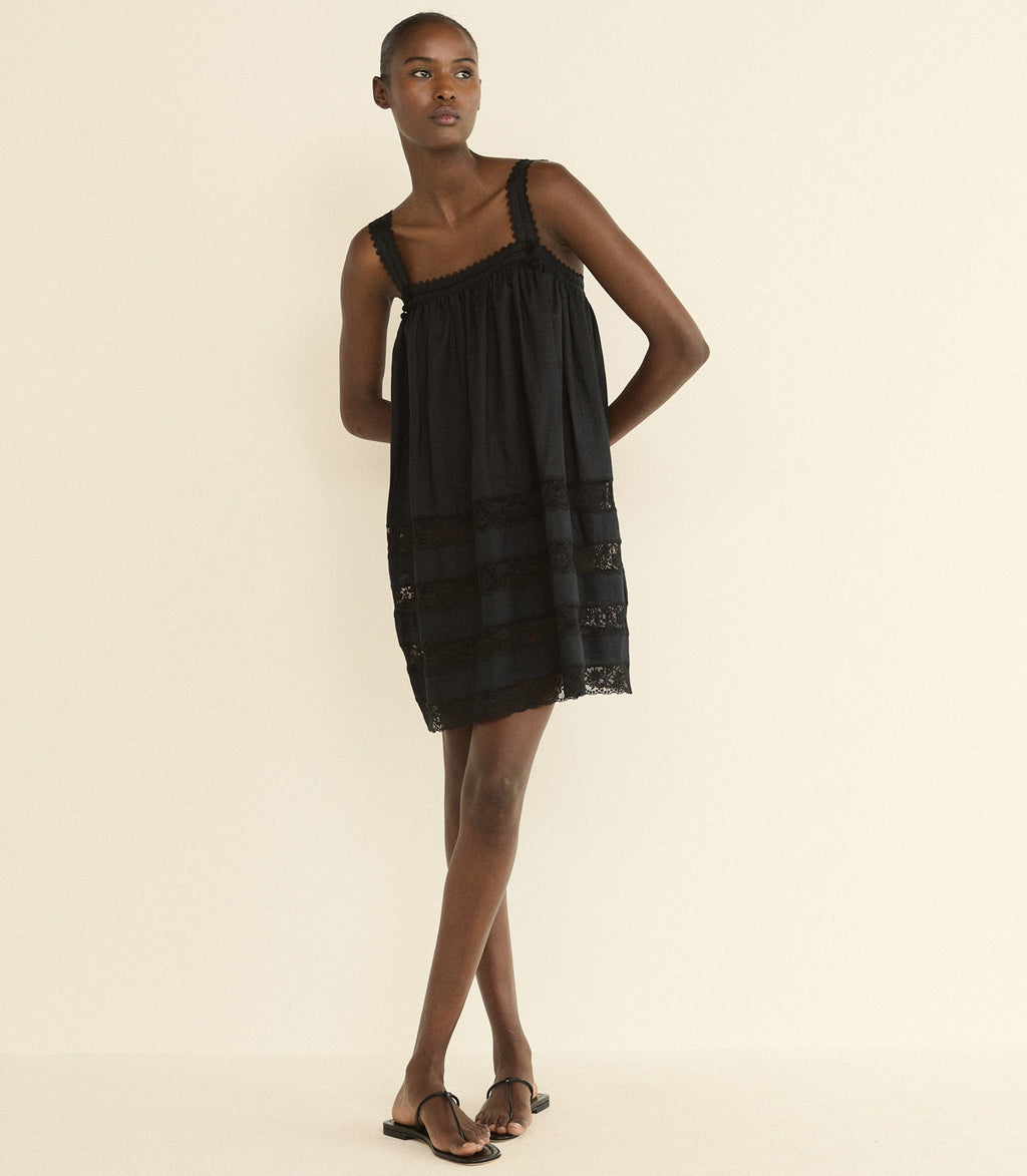 A model wearing a black sleeveless mini dress with a gathered bodice and sheer lace-like horizontal bands near the hem, paired with black sandals, against a plain cream background.