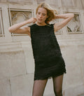 Woman wearing a black fringed top and skirt set standing in front of a stone wall.