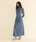 A woman modeling a long-sleeved, high-neck maxi dress with a blue, black, and white plaid pattern, standing against a solid beige studio background.