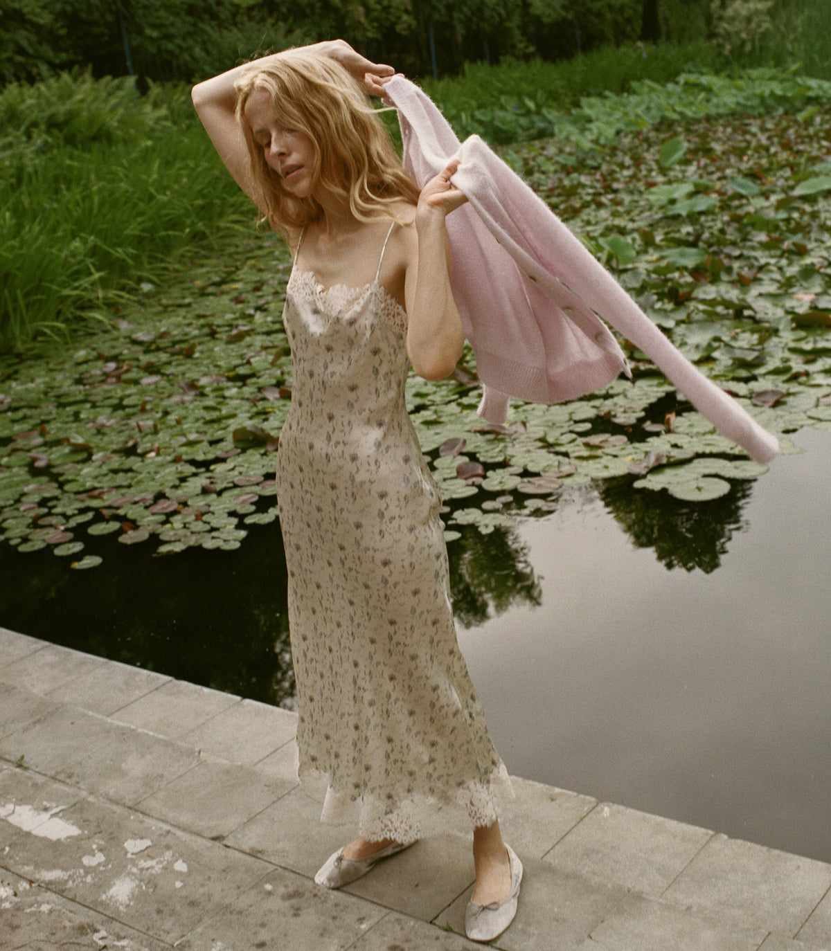 A woman stands on a stone walkway beside a pond with lily pads, wearing a light beige floral slip dress and holding a pale pink cardigan in front of ponds filled with lily pads.