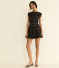 A woman modeling a short black dress with horizontal textured stripes, boat neck cap sleeves, and a flared skirt, paired with black heeled sandals against a plain cream background.