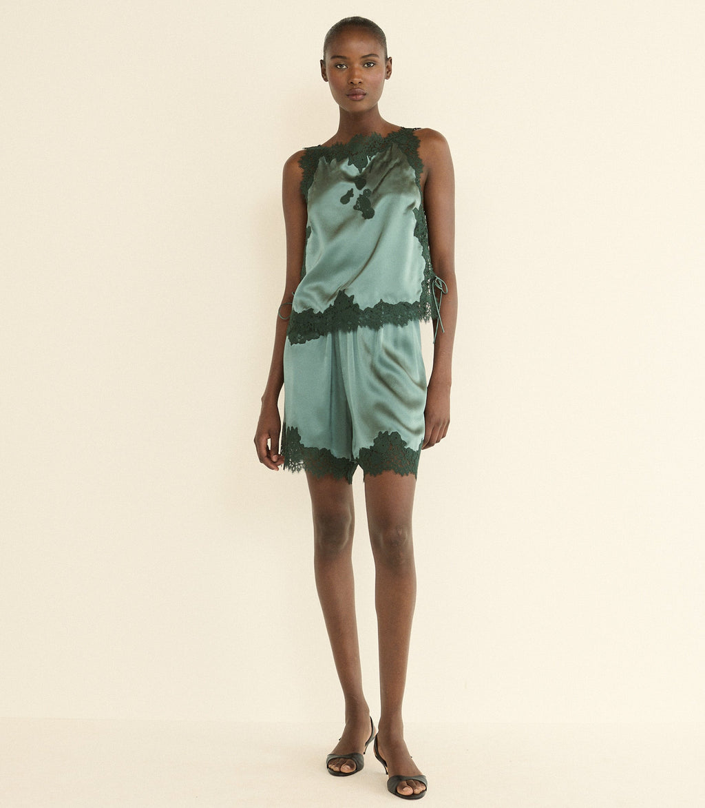 A model stands against a plain cream studio background wearing a sleeveless green satin top with dark green lace trim and a dark green abstract print.