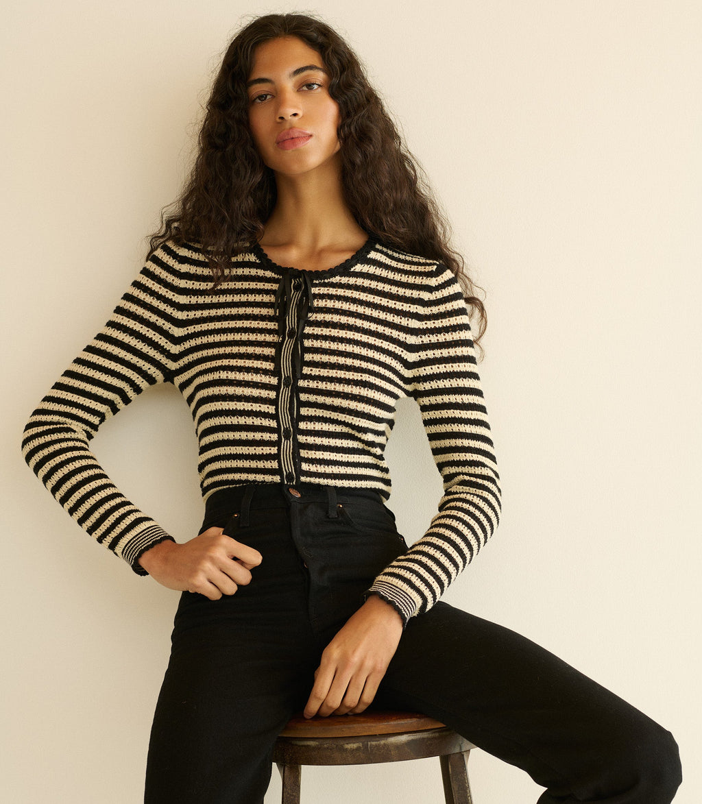 Woman wearing a black and white striped cardigan sitting on a stool against a plain background