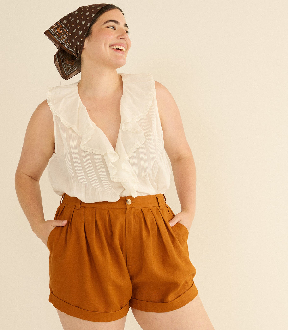 Woman wearing a white blouse and rust-colored shorts with a patterned headscarf on a beige background