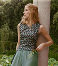 A woman in a sleeveless dark paisley top and sage green pants leans against a stone pillar in a lush garden setting.