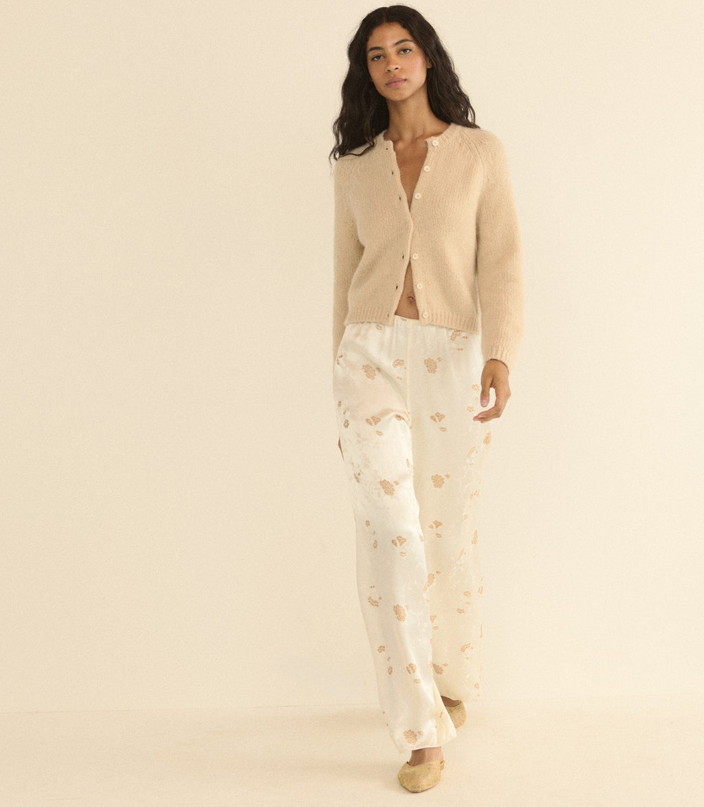 A woman modeling a cozy two-piece loungewear set consisting of a beige button-up cardigan and white pants with a light tan floral pattern, set against a solid cream background.