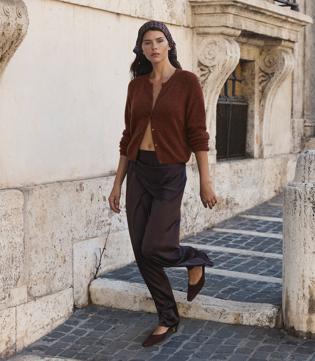 A woman wearing a rust-brown cardigan, dark brown draped trousers, and brown flats walks down a stone-paved street next to a beige stucco building.