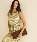 Woman wearing a patterned dress holding a reddish brown clutch against a beige background