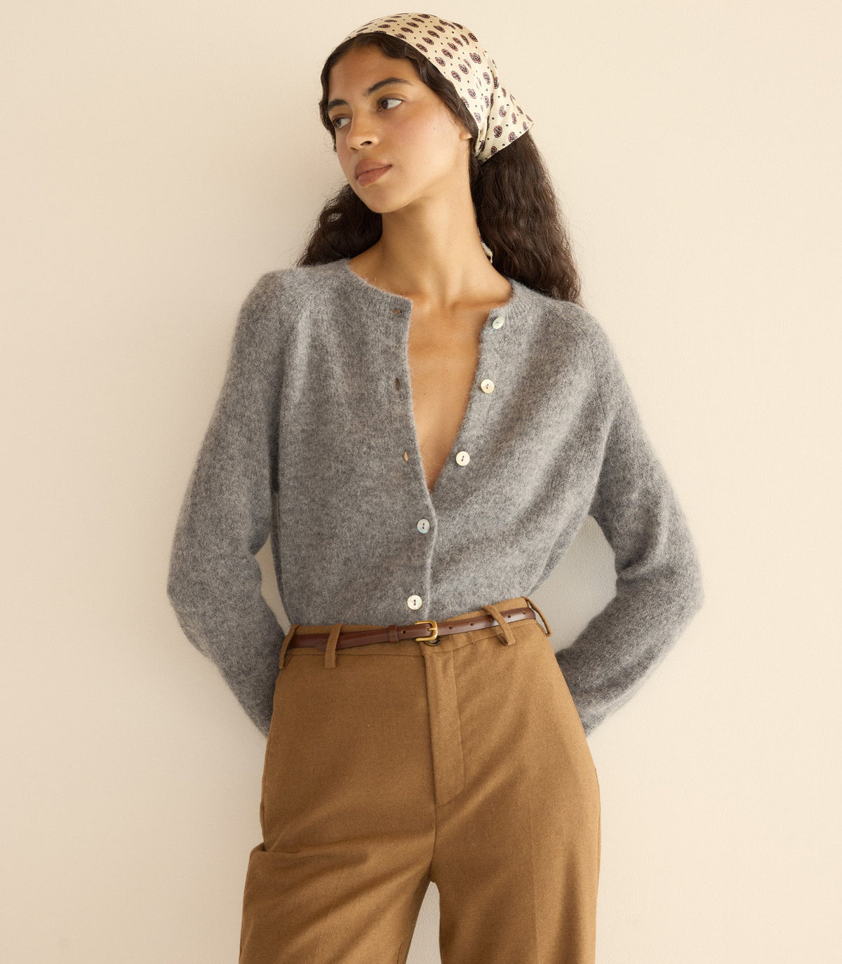 Woman wearing a gray cardigan and brown pants with a beige headband against a plain background