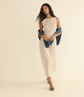 A woman modeling a sleeveless cream dress with a delicate floral print, styled with a draped blue shawl and metallic flat shoes, set against a solid warm beige studio background.