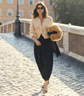 A woman walking on a cobblestone bridge wearing a beige knit cardigan, black wide-leg pants, and carrying a black coat with a fluffy tan fur trim.