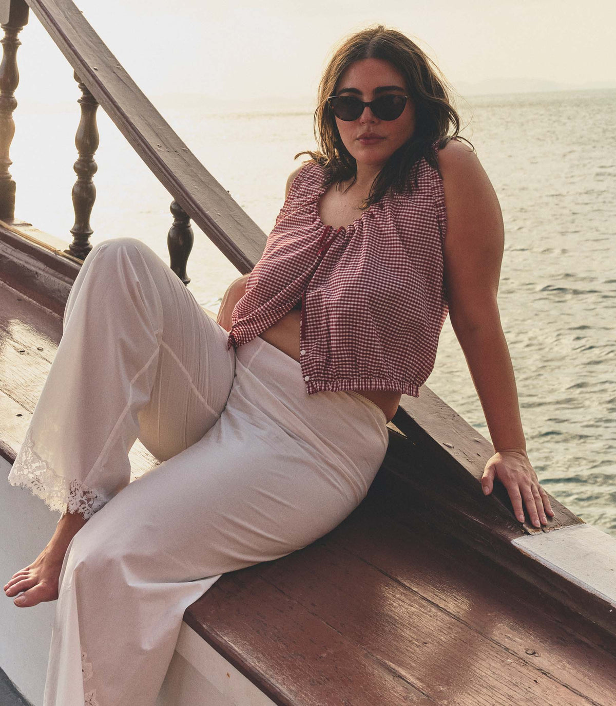 Woman sitting on a boat deck by the water wearing sunglasses and a red checkered top with white pants.