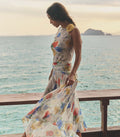 Woman in a white floral dress standing on a wooden platform by the ocean with mountains in the background.